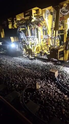 The video shows a large yellow railway maintenance machine operating at night on a gravel track. The machine has numerous hydraulic hoses and mechanical components. Several bright lights illuminate the work area. The machine appears to be working on the trackbed, with some of its lower components in contact with the ballast. The scene is dark, suggesting it is nighttime, and the lighting from the machine is the primary source of illumination. There are no visible people in the immediate vicinity of the machine's active work area.