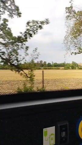 The video is shot from inside a vehicle, likely a bus or a car, looking out through the windshield. The foreground shows a dark dashboard with a USB port and a sticker with a charging symbol. Beyond that, a large field of dry, harvested crops is visible, with a thin wooden post standing in the middle of it. The background is a line of trees and a cloudy sky. On the right side of the frame, a large tree with green leaves partially obstructs the view of the field. The vehicle appears to be in motion, causing the scenery to move past.