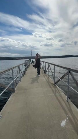 The video shows a woman with long hair, wearing jeans, a white top, a brown dress, white shoes, and glasses, walking on a concrete pier surrounded by water. The pier has metal railings. In the background, there is a body of water with land visible in the distance. The sky is blue with white clouds. The woman walks toward the camera, then stops and poses. There is a strong wind.