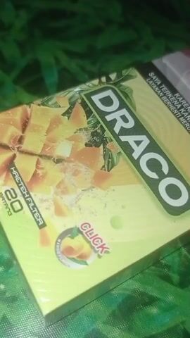 This is a close-up, static shot of a cigarette pack with a yellow and green design, featuring the brand name "DRACO" prominently displayed. Sliced mangoes are depicted on the front of the pack, along with a "CLICK" button graphic indicating a flavor capsule. The pack also shows "20 BATANG" and "HAZTEH FILTER" text. The background is blurred, showing a textured, dark green surface that appears to be fabric. The lighting is dim and focused on the cigarette pack, creating a slightly moody atmosphere. There are no people or actions depicted in the video.