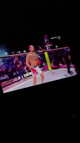 The video shows a man with tattoos on his chest and arms, wearing white shorts and red gloves, standing in a fighting ring. He is facing the camera and appears to be in the middle of a match or event. There are people in the background, some in the stands and others in the ring area, and the environment appears to be a sports arena with bright lighting.