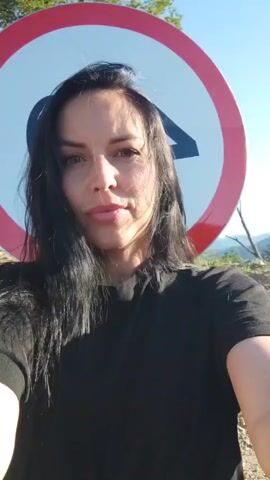 A woman with long, dark hair, wearing a black t-shirt, records a selfie video outdoors in Vardane-Verino, Russia. She stands in front of a large, circular speed limit sign displaying the number "40" in black on a white background with a red border. The sign is mounted on a metal post.

The woman looks directly at the camera, initially speaking with a neutral expression. She then puckers her lips into a playful "kissy face" or pout, holding the pose for the rest of the short clip, occasionally shifting her head slightly.

The setting appears to be a sunny day with a clear blue sky. The ground visible beneath the sign is dirt or gravel. In the background, distant hills and sparse greenery are discernible. A small, partially visible green sign is situated below the main "40" sign, though its text is unreadable.