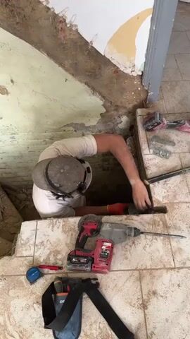 The video shows a person in a white t-shirt and a cap working in a hole in the floor. They are wearing gloves and appear to be using a tool to break up concrete or tile. Several power tools, including a circular saw and a hammer drill, are visible on the floor nearby, along with a measuring tape and a tool belt. The environment appears to be a construction or renovation site, with exposed brick and plaster walls.