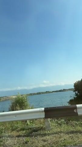 The video shows a roadside view of a body of water with a clear blue sky above. The foreground features a guardrail with white and black reflective strips, and dry grass and dirt. Behind the guardrail, a body of water is visible, with some vegetation and distant hills or mountains. The camera appears to be moving, as the perspective shifts slightly throughout the short clip. There are no people or animals visible in the frame. The environment appears to be a natural landscape, possibly a lake or reservoir, with evidence of a road and its accompanying infrastructure. The lighting suggests it is daytime.