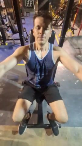 A man is working out on a butterfly machine in a gym. He is seated and grasping the handles of the machine. He moves his arms to bring the handles together in front of his chest, then slowly returns them to the starting position. He is wearing a blue tank top, black shorts, and black sneakers. The gym has various other exercise machines and weights visible in the background. The floor is concrete, and the lighting is somewhat dim.