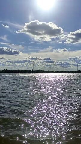 The media file captures a serene waterscape with a solitary jet ski rider in the distance. The sun is low in the sky, casting a shimmering, sparkling reflection across the water's surface, suggesting it is either early morning or late afternoon. The sky is dramatic, with a mix of bright sunlight and dark, textured clouds. A silhouetted treeline forms the distant horizon, with a tall, slender structure, possibly a radio tower or pylon, visible on the left side. The overall impression is one of peaceful solitude and enjoyment of a natural setting. The jet ski, a recreational watercraft, is the only human-made object clearly in motion, carving a white wake through the water. The scene conveys a sense of freedom and a connection with nature. There is no visible text in the media file.