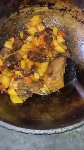 A top-down, close-up shot shows food being cooked in a dark, round pan. The pan contains chunks of potatoes, carrots, and meat, all appearing to be sauteed in oil. A black spatula is visible, stirring the ingredients. The background is blurry, suggesting a simple cooking environment. The lighting is dim, with some highlights reflecting off the food and pan.
