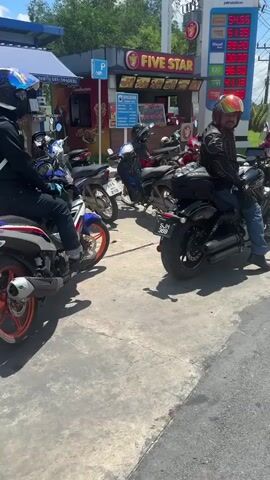 Two individuals on motorcycles are stopped at a gas station. The person on the left is wearing a blue helmet and a black jacket with an Oakley backpack. Their motorcycle is white, red, and blue. The person on the right is wearing a colorful helmet and a black jacket, and is on a black motorcycle. They are looking towards the right. In the background, there are other motorcycles parked, a "FIVE STAR" convenience store, a gas price display showing fuel costs in Thai Baht, and lush green hills under a partly cloudy sky. Power lines and poles are visible above the scene.