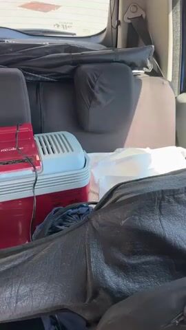 The video shows the interior of a vehicle, likely the back seat or cargo area. Various items are packed in, including bags, blankets, and a red and white cooler with a red electronic device on top. A person's hand with pink nail polish is visible in the foreground, resting on a black car seat cover. The background appears to be the outdoors, seen through the vehicle's rear window.