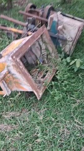 In an outdoor scene, there is a patch of grass scattered with various pieces of old, rusty machinery. The equipment is heavily weathered and appears to be parts of larger devices, possibly engines or industrial equipment. The grass is green and unkempt. The scene conveys a sense of disuse and abandonment of the machinery.