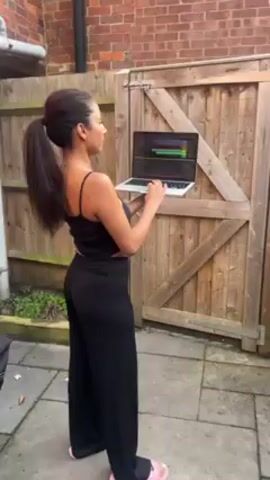 A woman stands outside in a yard holding a laptop. She wears a black tank top and pants. Her hair is pulled back into a ponytail. She looks at the laptop screen and types on the keyboard. The laptop screen displays what appears to be an audio or video editing program. Behind her is a wooden fence and a brick building. The ground is paved.