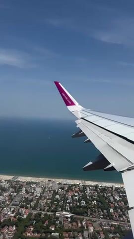 The video shows an aerial view from an airplane window. The camera is pointed downwards and slightly to the side, capturing the airplane's wing in the foreground. The wing is white with a purple tip that has the Wizz Air logo. Below the wing, a coastal city is visible, with buildings densely packed together and trees interspersed throughout. A beach area separates the city from the ocean, which is a deep blue. The sky above is clear and blue with a few wispy clouds. The perspective suggests the plane is either approaching or departing from an airport near the coast.