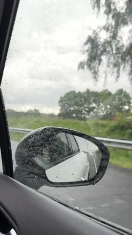 The video, shot from the interior of a car, shows a rainy highway drive. Raindrops streak the side window and cover the rearview mirror. As the perspective shifts towards the windshield, operating windshield wipers clear the rain, revealing other vehicles on the road ahead, blurred by the rain. The sky is overcast and gray.