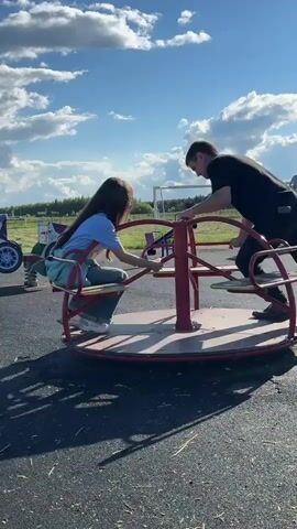 This video captures a heartwarming moment on a playground in Mursalimkino, Russia. The primary subjects are a man and a young girl. The man, wearing a black t-shirt and dark pants, is actively engaged in spinning a merry-go-round. The girl, with long dark hair and wearing a light blue shirt and jeans, sits on one of the merry-go-round's seats, her face obscured by her hair, suggesting she's enjoying the ride. The man is positioned outside the merry-go-round, leaning in to provide the momentum.

The setting is an outdoor playground with an asphalt surface. The merry-go-round itself is a classic metal structure with red supports and wooden seats. In the background, other playground equipment is visible, including what appear to be spring riders shaped like vehicles. Beyond the playground, a flat, open landscape stretches out, dotted with distant trees or structures and a fence line. The sky is a prominent feature, filled with fluffy white clouds against a bright blue backdrop, indicating it's daytime, likely late afternoon due to the long shadows cast on the ground. The overall atmosphere appears joyful and relaxed, as the man and girl share a simple, fun activity. No visible text is present in the video.