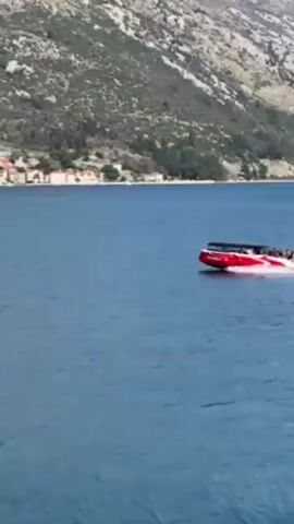 A red speedboat with a black roof moves quickly across a blue body of water, leaving a white wake behind it. The boat appears to carry passengers. In the background, a mountainside with a town or village is visible along the shoreline. The sky is not visible in the shot.