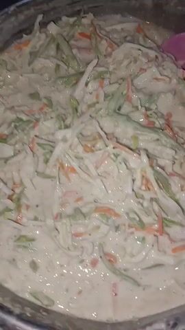 A close-up shot shows a person's hands stirring a creamy mixture of shredded vegetables, possibly coleslaw or fritters, in a large silver bowl with a pink utensil. The vegetables appear to be primarily cabbage and carrots. The mixture is thick and appears to be a batter or dressing coating the vegetables. The video is grainy and low-light, with the focus primarily on the food and the stirring action.