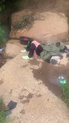 The video shows a pile of laundry on a large rock next to a body of water. A person is lying on the rock, covered by a green jacket and surrounded by clothes. A black basin filled with clothes is also visible on the rock. The person is wearing white clogs. The scene appears to be outdoors, with vegetation and more rocks in the background. The water is murky.
