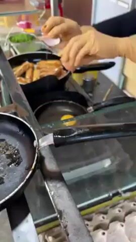 The scene shows a street food vendor preparing food. A person in a hijab is working behind a metal cart with a glass top. The cart has a two-burner gas stove, on which two pans are placed. One pan contains several sausages, and the other contains a single fried egg. The person cracks another egg into a small white container. They then add a white liquid from a small silver container into the pan with the egg. There are squeeze bottles of condiments on the counter, along with containers of leafy greens.