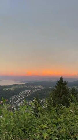 The video shows a wide landscape view at sunset. The sky is a gradient of orange, pink, and gray hues. Below the sky, there are orange and pink clouds, illuminated by the setting sun. In the distance, there is a body of water and a hazy outline of mountains. Closer in, there is a town or city with buildings nestled among rolling green hills and dense forests. In the foreground, there are green bushes and the top of a dark green conifer tree. The video is shot from a high vantage point, looking down on the landscape. There are no people or animals visible in the video.
