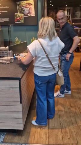 The video shows a woman in a white sweater and jeans approaching a counter in what appears to be a cafe or bakery. She is facing away from the camera and towards a person behind the counter, who is wearing a red apron and black shirt. The counter is made of dark material and has wooden paneling on the front. There are coffee cups, sugar dispensers, and a napkin holder on the counter. In the background, there is a menu board with pictures of coffee and coffee beans, as well as other customers standing at the counter. The woman appears to be interacting with the person behind the counter, possibly placing an order. The environment appears to be indoors, possibly in a shopping mall or a public space, with tiled floors and other shops visible in the background. Another customer, a woman in a pink and white striped shirt, is also visible at the counter, interacting with another employee. Further in the background, a man is sitting on a bench with a young child.