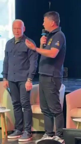 Two men are standing next to each other on a stage. The man on the left has short white hair and is wearing a dark blue button-down shirt, black pants, and dark sneakers. He is holding a small black object in his left hand. The man on the right is holding a microphone and speaking. He is wearing a black polo shirt with a yellow and blue emblem on the chest, black pants, and dark sneakers. A cream-colored chair is visible in the foreground. A screen with what appears to be the letters "МГ" is in the background. The two men appear to be engaged in a conversation.