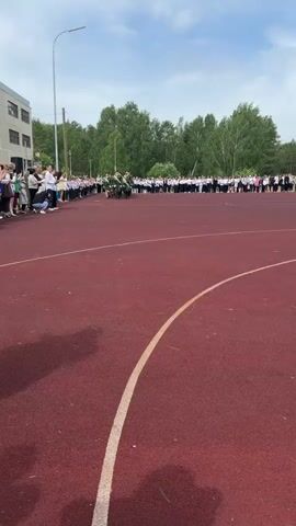 The video shows a group of young people in green uniforms marching on a red athletic field, in front of a larger crowd of spectators.  The marchers are in a formation and are saluting as they move forward.  The field has white lines on it.  In the background is a building and trees, under a partly cloudy sky.