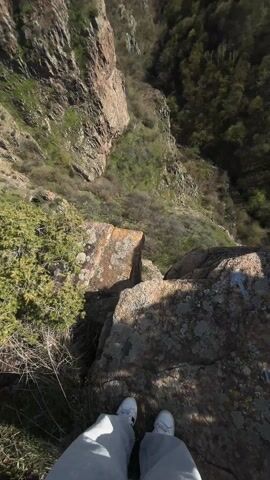 The camera, positioned on a rocky precipice, slowly pans upwards to reveal a deep, wooded valley. The foreground is dominated by moss-covered rocks and a small, green bush. A pair of white shoes are visible at the bottom of the frame, indicating the location of the camera holder. The valley is flanked by steep, rocky cliffs on one side, while the other side is covered with dense forest. In the distance, rolling hills and a clear, blue sky with a few scattered clouds complete the landscape. The overall impression is one of a vast, natural environment viewed from a high vantage point.