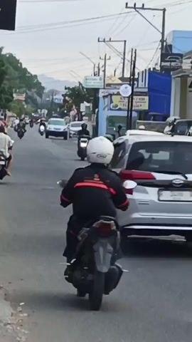 The video shows a street scene with several motorcycles and cars. A motorcycle with two people on it is in the foreground, facing away from the camera. The person in the back is wearing a black jacket and blue shorts, and the person in front is wearing a black helmet. To the left, another motorcycle with one rider is also moving away from the camera. In the background, there are more motorcycles and cars on the road, as well as buildings and utility poles. The sky is overcast. The video appears to be shot from a moving vehicle.