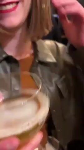 A woman is holding a glass of a creamy, frothy drink and smiling broadly at the camera. She is wearing a brown leather jacket over an orange top, and has short brown hair and bright red lipstick. Her teeth are visible as she laughs. In the background, another person is standing at a bar, wearing a black shirt and light-colored pants. The setting appears to be inside a bar or restaurant, with a blurred background of other people and interior elements. The woman seems to be enjoying herself.