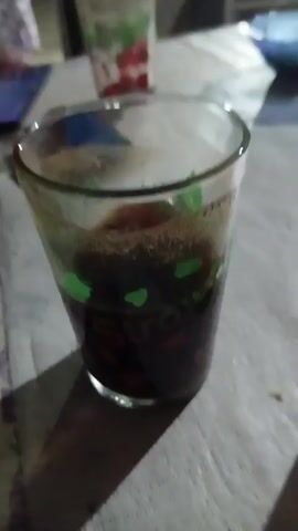 The video shows a close-up of a glass of dark liquid, possibly coffee or soda, with bubbles. The glass has a decorative pattern of green hearts and red fruit. In the background, another glass with a similar pattern is visible, along with a smartphone lying on a table covered with a plastic tablecloth. Further in the background, there are shelves filled with items, a blue bowl, and a person in a black hijab and patterned skirt walking away from the camera. The scene appears to be an outdoor or semi-outdoor establishment, possibly a food stall or small shop, with dim lighting and a rustic atmosphere.