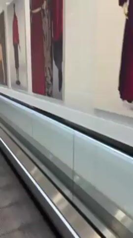 The video shows a moving walkway with artwork lining the walls. The walkway is made of silver metal and has a white barrier along the side. The artwork consists of various fashion images, including dresses, figures, and accessories. The lighting in the area is bright and even. The walkway is moving, taking the viewer past the artwork. The overall scene appears to be within an airport or shopping mall setting.