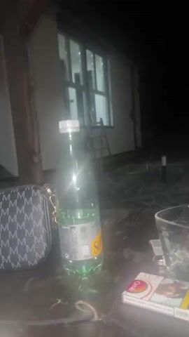 The video displays a static, dimly lit outdoor scene at night. In the foreground, on a dark table surface, there is a partially visible black and white patterned bag, a green glass bottle with a yellow label (Schweppes) containing a clear liquid, a clear drinking glass, and a pack of white, red, and yellow cigarettes with an image on it, upon which a small lighter rests. In the midground, a building with illuminated windows and a ladder leaning against it is visible. The background is dark and indistinct. No people or specific actions are depicted.