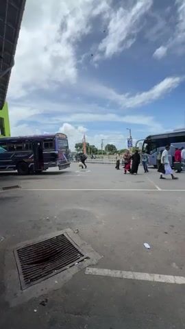 The video captures a dynamic daytime scene at what appears to be a bus terminal or a busy public transport area in Hambantota, Sri Lanka. The sky is bright blue with scattered white clouds, indicating sunny weather.

Several buses are visible across the paved area. On the left, a purple bus is parked. In the center, a large, dark blue bus is stopped, with people walking towards and away from its open door, some seemingly boarding or disembarking. Further down the street, a light blue bus with decorative patterns is parked, and a red bus is partially visible on the far right.

Numerous people are moving across the scene. They are seen walking, some carrying bags, and interacting with the buses. Some individuals are dressed in what appears to be traditional attire, including long garments and sarongs. In the background, other vehicles like a white van and motorcycles are present, alongside distant structures and utility poles with overhead wires.

The ground is asphalt, marked with white lines, and a metal storm drain cover is prominent in the foreground. Some scattered litter is visible on the pavement. The overall impression is one of everyday activity at a bustling transport hub, with people going about their daily routines. No specific text on signs or vehicles is clearly discernible.