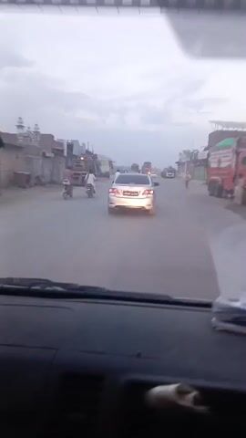 The video shows a car's dashboard view while driving on a road. Several other vehicles, including cars, trucks, and motorcycles, are visible in front. Buildings line both sides of the road, which is dusty and appears to be in a less developed area. The sky is overcast. The sound of a siren is prominent throughout the video.