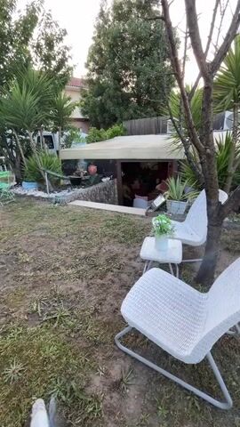 The video shows a small, sloped backyard with two white, woven plastic chairs and a small white table holding a pot of artificial flowers. The ground is mostly dirt and patchy grass. In the background, there's a covered patio-like structure built into the slope of the yard, with glimpses of people inside. Beyond the yard, the terrain rises to show houses and vegetation against a blue sky. The camera pans around the backyard, across the landscape, then tilts upward to the sky.