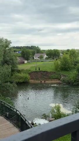 A group of people are near a river. The riverbank is steep and covered in vegetation. In the foreground, the river water is choppy. On the riverbank, two young men are standing in the shallow water. Further up the bank, on top of the rise, three figures stand near a utility pole. The background shows houses and trees under a cloudy sky.