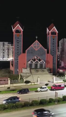 A nighttime shot shows a church with two tall towers, each topped with a cross. The church is lit up and has a large set of steps leading to three doors. A street runs in front of the church, with several parked cars. Cars drive along the road. Apartment buildings are visible in the background. Bells ring at the beginning of the video.