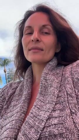A woman with brown hair and brown eyes, wearing a grey knitted cardigan, is filmed outdoors in a selfie style video. She is smiling and looking at the camera. Behind her, there is a splash pad with blue and red surfaces, mushroom-shaped water features, palm trees, and a bright blue sky with white clouds. There are power lines and a building visible in the background. The video is short and seems to be a brief update or greeting.