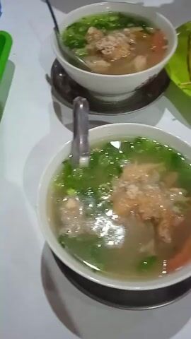 The video displays a white table upon which sits two bowls of clear soup, topped with green onions and small, unidentified brown solids. Each bowl contains a silver spoon and rests on a dark-colored saucer. Adjacent to the soup is a small green plate, containing a fried item and a skewer holding three small, round, brownish food items. A partially visible plastic bottle of water is also on the table. The scene is brightly lit, and the camera is positioned directly above the table, providing an overhead shot of the food items.