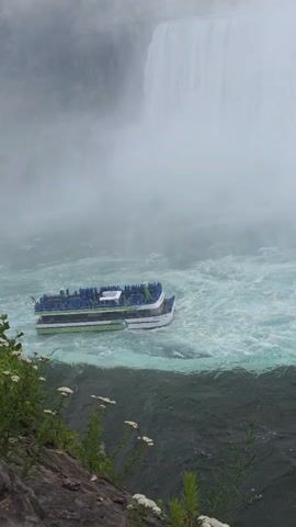 A tour boat with people on its deck navigates through turbulent, misty waters near the base of a large waterfall. The boat is green and white with blue seats. The water is a mix of dark green and turquoise, churned by the waterfall's powerful cascade, which is largely obscured by mist. Rocky terrain covered in green foliage and small white flowers is visible in the foreground on the left side of the frame.