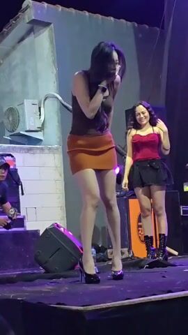 The video shows two young women on a stage. The woman in the foreground is taller and wearing a brown tank top and an orange skirt, with black high heels. She is running her hand through her hair and looking to her right. The woman in the background is shorter and wearing a red top and a black skirt with black boots. She is smiling and looking at the camera. There is stage equipment behind them, including speakers and lights. The environment appears to be an indoor venue, possibly a club or concert hall.
