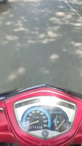 The video is filmed from the first-person perspective of someone riding a red motorcycle on a paved road. In the distance, several yellow dump trucks are seen approaching and passing the motorcycle. On the right side of the road, there are trees and what appear to be small shops or houses. Another person on a white motorcycle is also seen driving in the opposite direction.