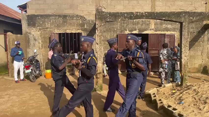 This media file captures a scene in Gudi, Nigeria, likely a training exercise or drill involving young people in uniform. The central figures are several young men dressed in navy blue uniforms with yellow trim, wearing matching caps. They are holding what appear to be rifles, suggesting a cadet or paramilitary group. They are positioned in a staggered formation, moving forward, with some actively engaged in what seems like marching or drill practice.

In the background, other individuals are present. To the left, a man in a light blue shirt and white pants stands near a parked motorcycle. Further back, a man in a pink shirt observes the activity. A few other figures are visible near doorways of a building constructed of rough, grey cinder blocks. The building has barred windows and wooden doors. The ground is a mixture of dirt and sand, with some scattered debris. A pile of concrete blocks is visible on the right side of the frame.

The lighting suggests it is daytime, with sunlight casting distinct shadows, indicating a clear or partly cloudy sky. The overall atmosphere appears serious, focused on the task at hand for the uniformed individuals. There are no visible signs or text within the frame. The scene is taking place outdoors in what seems to be a semi-urban or rural setting, given the building style and unpaved ground.