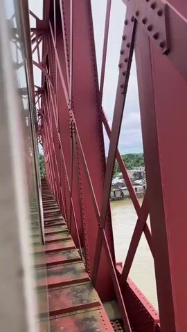 The video shows a view from a moving train crossing a bridge over a body of water. The bridge is made of red metal girders and rivets. The train cars are partially visible on the left side of the frame, and the tracks are visible below. The water is murky brown, and there are trees and buildings on the far bank. The sky is overcast. The video appears to be filmed on a slightly windy day.
