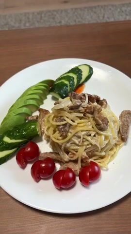 A close-up shot shows a white plate with a meal on a wooden table. The meal consists of pasta with a creamy sauce and chunks of meat or mushrooms. On the side of the plate, there are sliced avocados, sliced cucumbers, and cherry tomatoes. The camera then pans out to reveal a television screen displaying a scene with a person wearing a red top. The television is mounted on a wooden cabinet with several drawers. A guitar is visible to the left of the cabinet. The floor is wooden, with a carpeted area in the foreground.