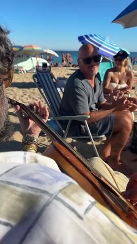 A first-person perspective video captures a close-up of a person's hands playing an acoustic guitar on a sunny beach. In the background, a bald man wearing sunglasses and a gray t-shirt sits in a striped beach chair, smiling and appearing to sing along. Beside him, a blonde woman in a white bikini top and a New York Yankees baseball cap also smiles and claps her hands. They are seated on the sand, with other beachgoers and umbrellas visible further in the distance. The ocean and a clear blue sky are visible at the top of the frame. The song appears to be in Spanish, with lyrics like "duse, durase fragancia de su corpo" audible.