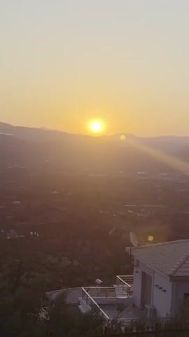 The video shows a landscape at sunset. The sun is low in the sky, casting long shadows and a warm glow over rolling hills and a body of water. In the foreground, there is a house with a tiled roof and a balcony. There is also some vegetation, including trees and bushes. The overall scene is peaceful and serene.