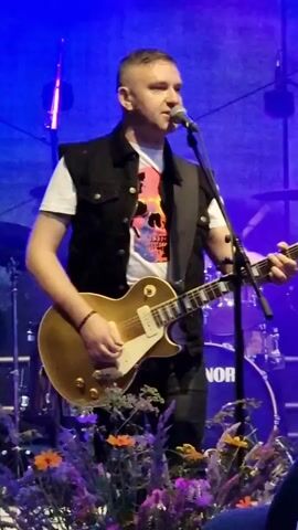 A man with short, dark hair and a shaved head is playing an electric guitar on a stage. He is wearing a white t-shirt with a colorful skull graphic, a black sleeveless vest, and dark pants. He is holding a gold-colored electric guitar and strumming it. A microphone is positioned in front of him. In the background, a drum set with a "SONOR" logo is visible, along with stage lighting and a blue backdrop. In the foreground, there are flowers and some audience members. The video is a static shot of this scene.