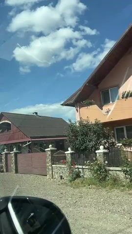 A video clip shows a car driving by houses and rural scenery. The sky is blue with white clouds. The houses vary in architectural style, from traditional wooden homes to more modern buildings with fences and decorative elements. There are some agricultural vehicles and equipment visible. The road appears to be unpaved or gravel.