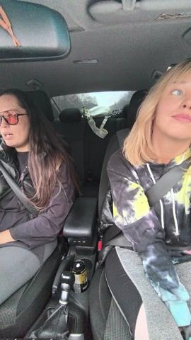 The video shows two women in the front seats of a car. The woman on the left has long, dark hair, is wearing sunglasses, a black vest, and gray pants. The woman on the right has blonde hair, a tie-dye hoodie, and gray pants. Both are wearing seatbelts. The car appears to be moving, and loud music is playing. There is a doll hanging from the rearview mirror and a drink in the console.
